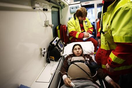 Raskaana oleva romaninainen makaa ambulanssin sängyllä. Hänen ympärillään on kaksi keltapunaisiin asuihin pukeutunutta ensihoitajaa. 