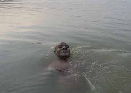 Tummaihoinen mies kelluu vedessä. Miehestä on vedenpinnan yläpuolella ainoastaan kasvot. Miehellä on silmät kiinni 