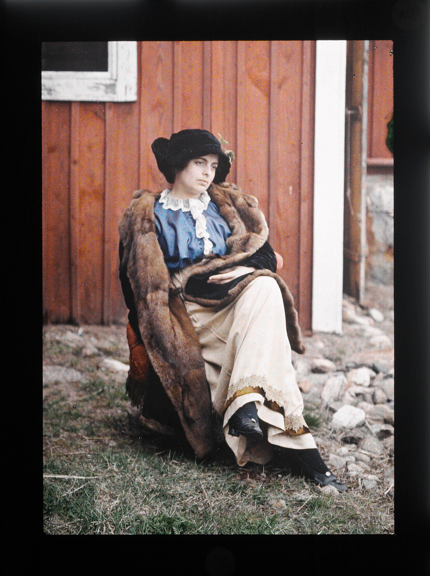 A lady sits pensively in front of a wooden building with a fur coat over her shoulders.